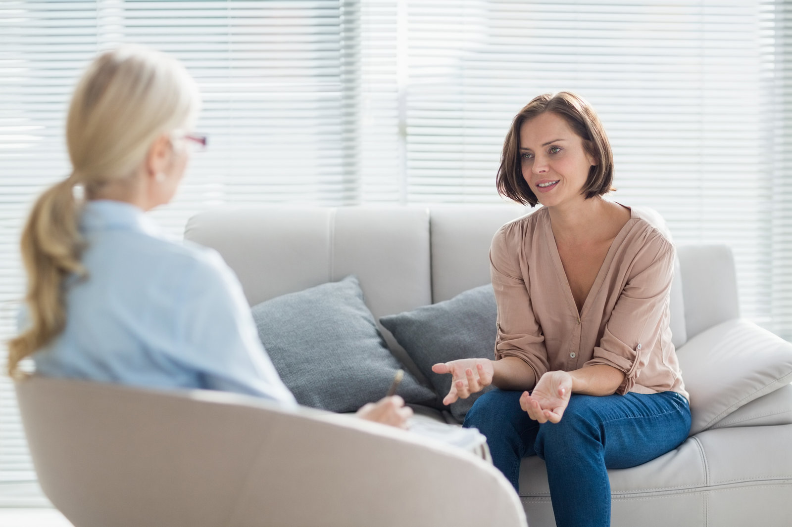 A woman sitting on a sofa talking with her hands open, while a therapist opposite takes notes