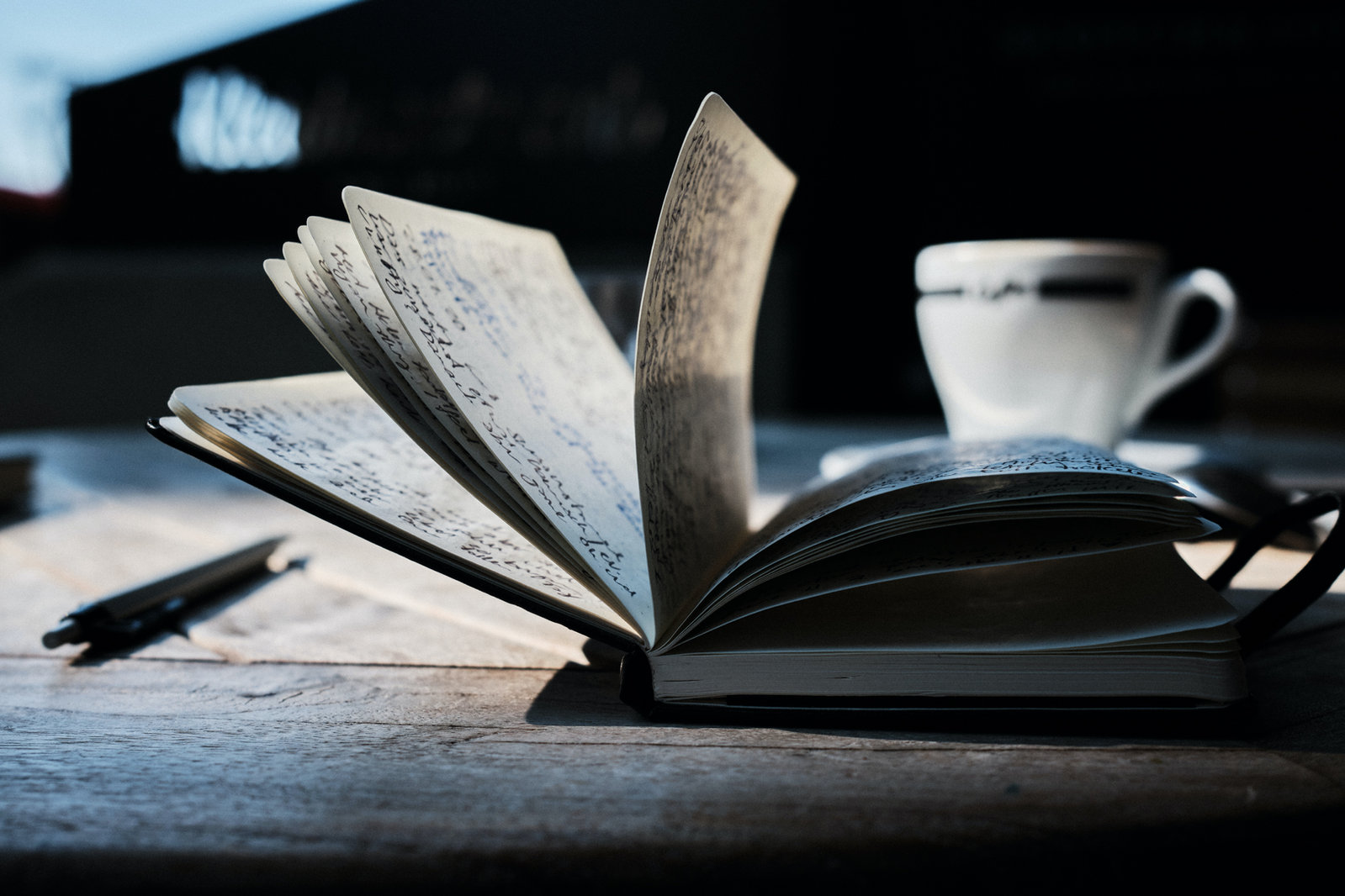An open handwritten notebook on a wooden table with a pen and a cup of coffee
