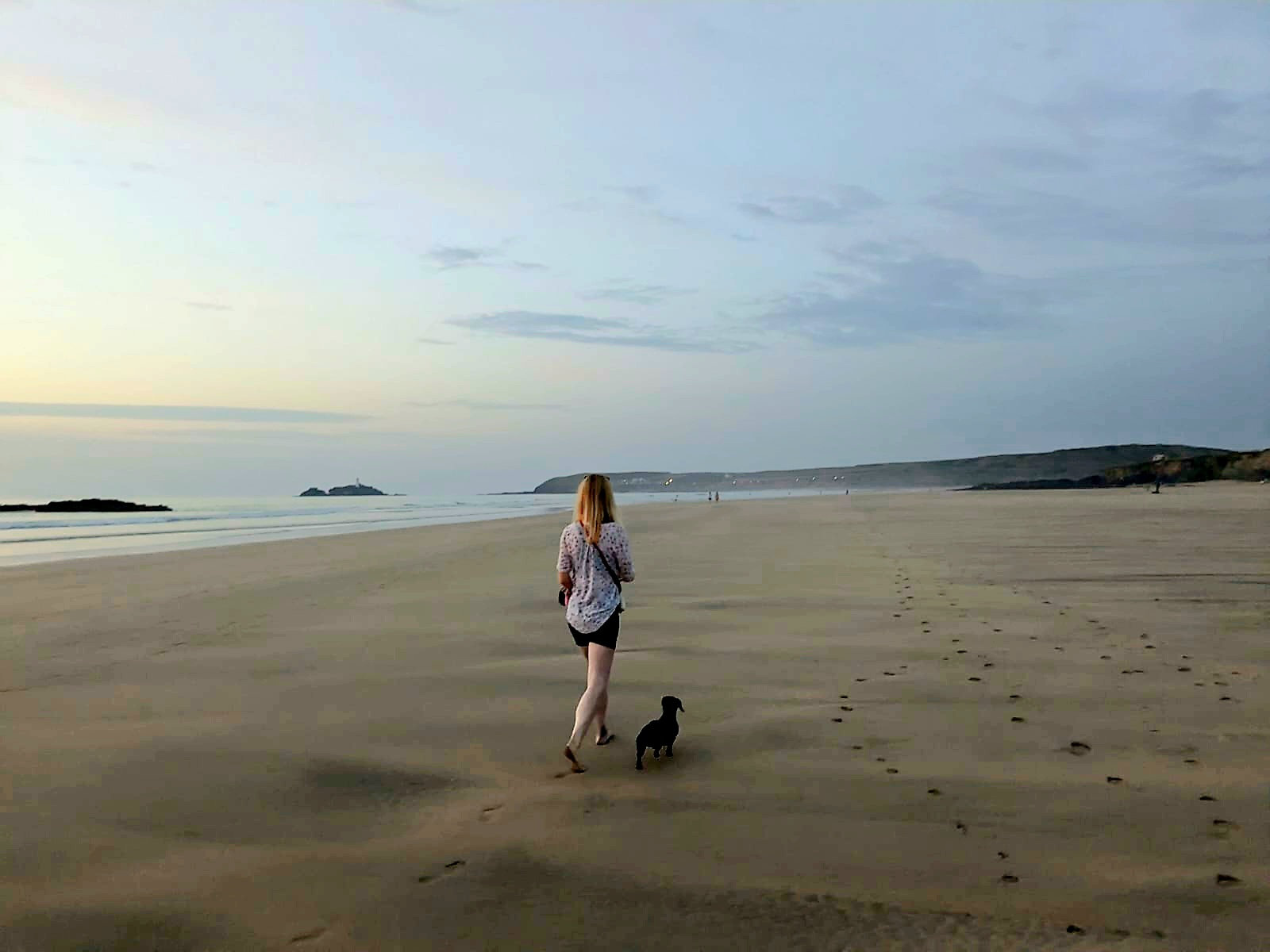 A woman walking a small dog along a wide empty beach at dusk