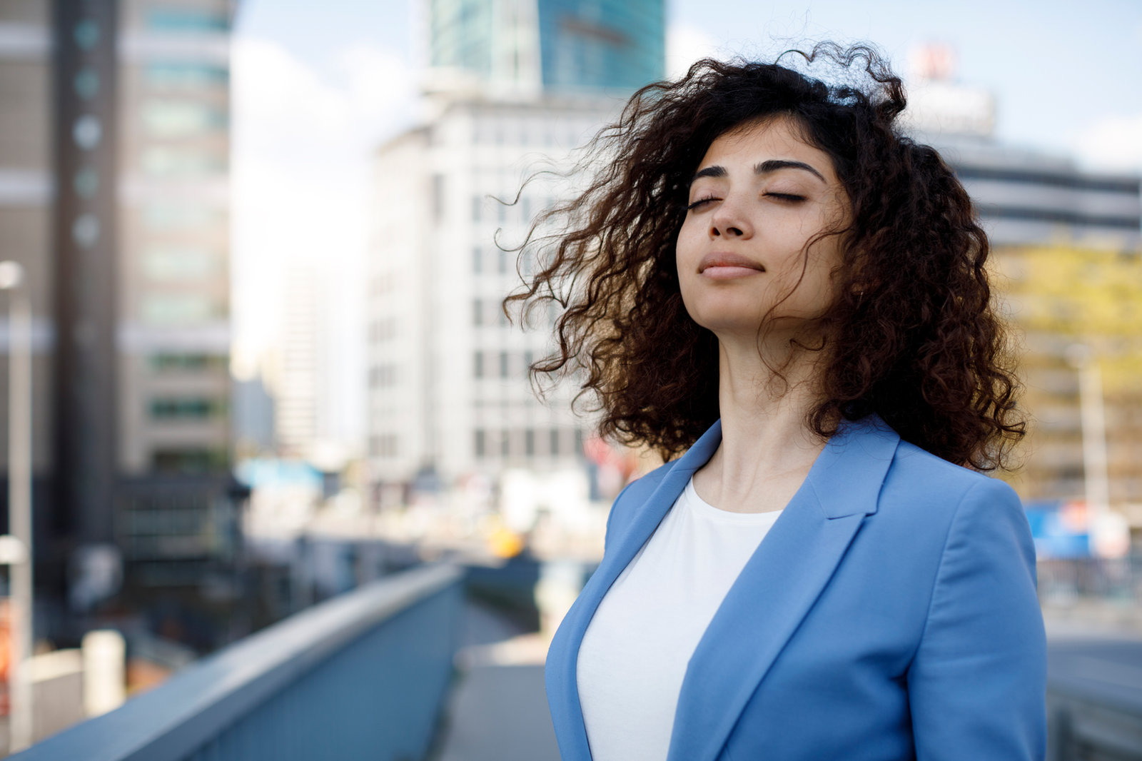 A woman standing outdoors in a city with her eyes closed, taking a slow breath