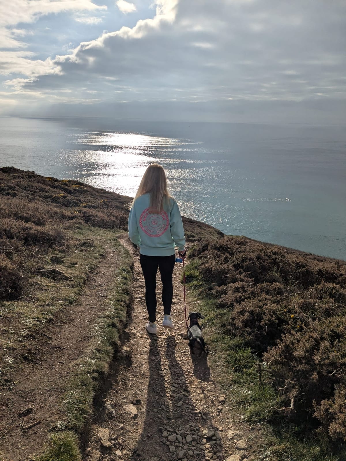 A woman walking a dog along a coastal path high above a sunlit sea