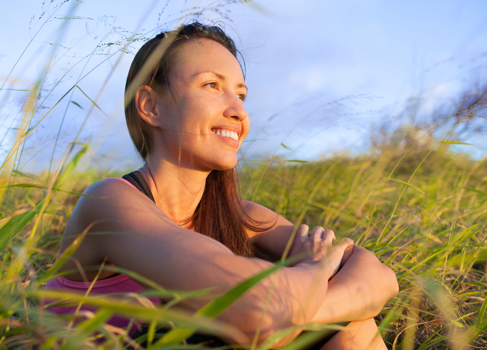 A woman sitting outdoors in long grass at golden hour, smiling and looking into the distance