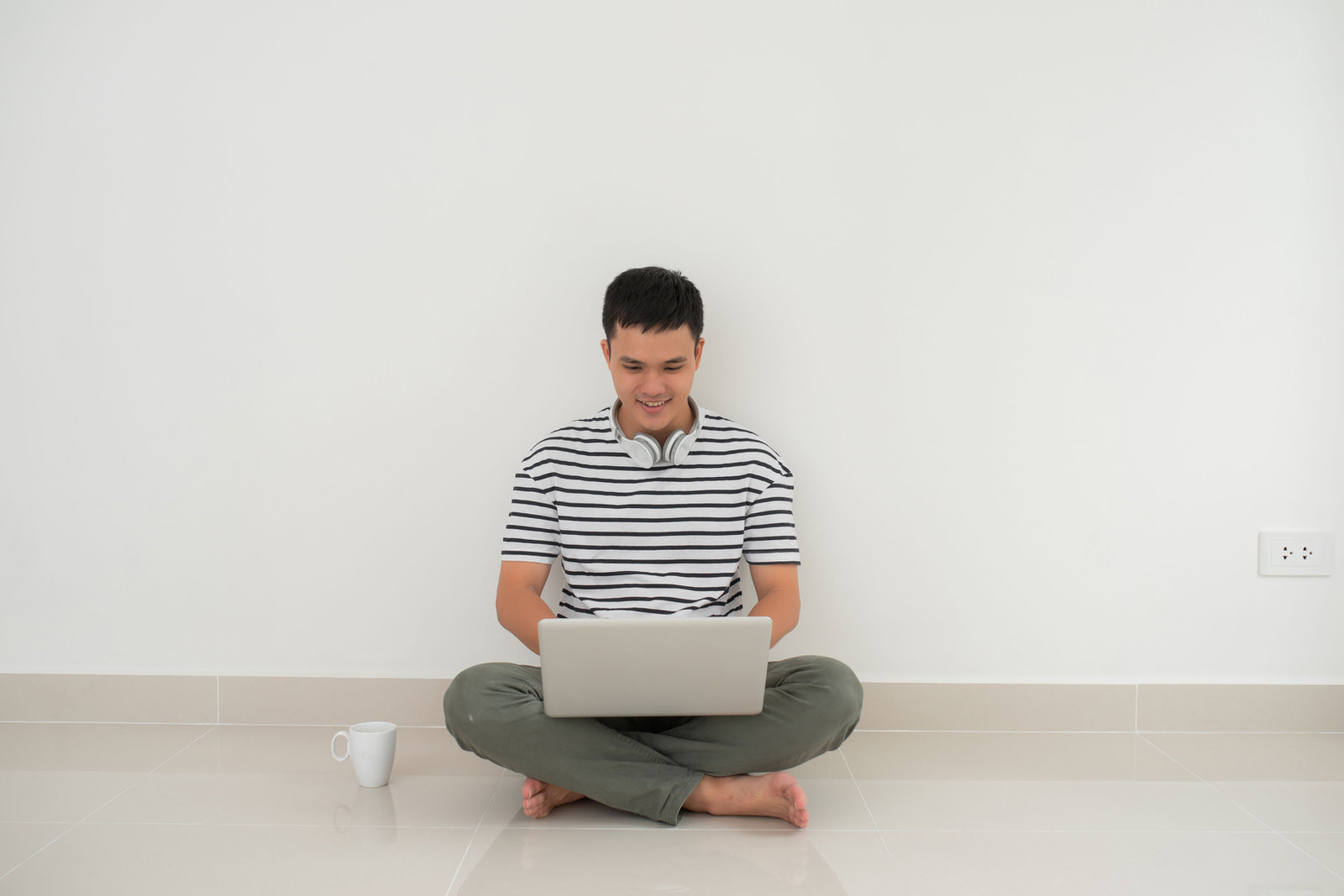 A man sitting cross-legged on the floor with a laptop, headphones around his neck and a mug beside him