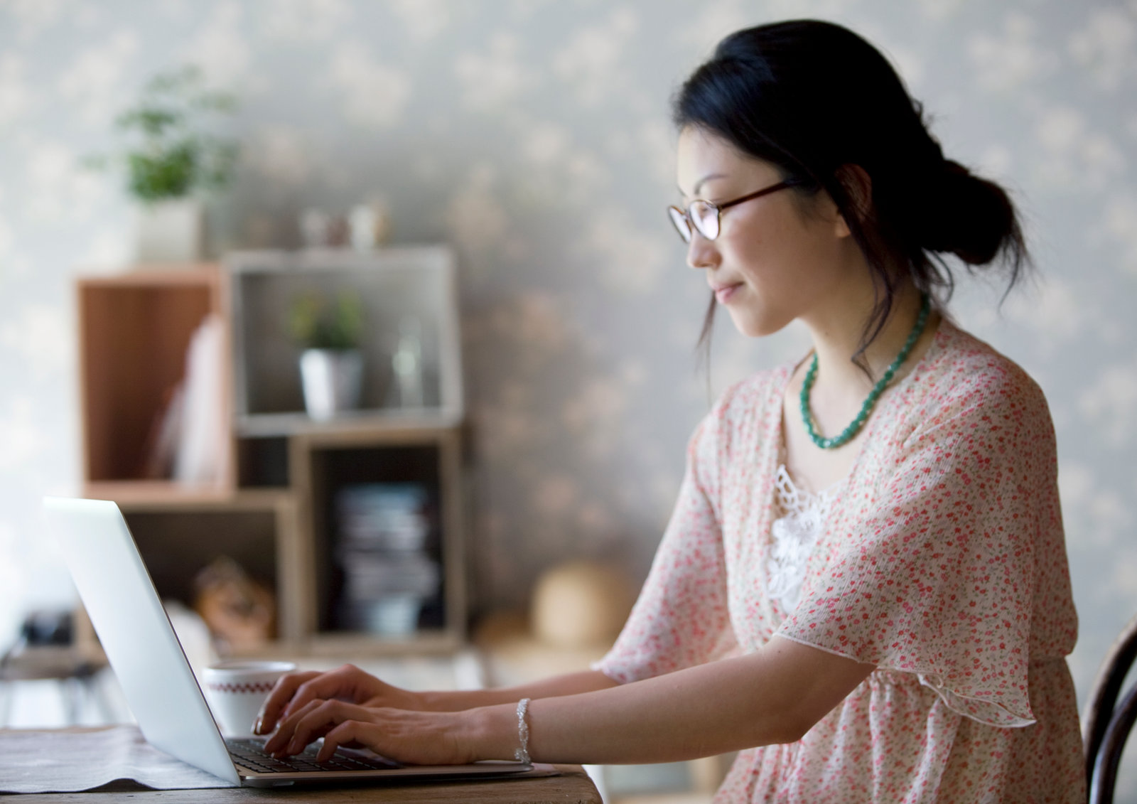 A woman typing at a laptop at a table at home, a mug beside her