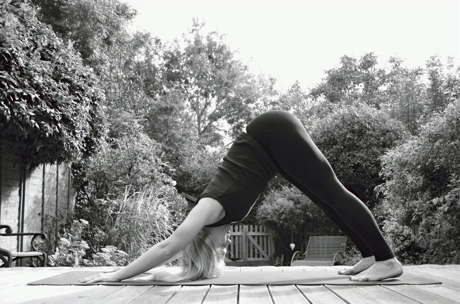 Black and white photograph of a woman in downward facing dog on a yoga mat on garden decking
