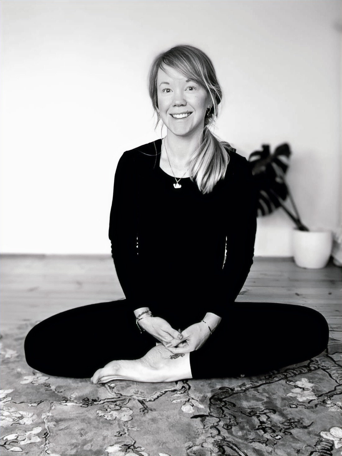 Black and white photograph of Dr Kayleigh Darch sitting cross-legged on a patterned rug, smiling at the camera