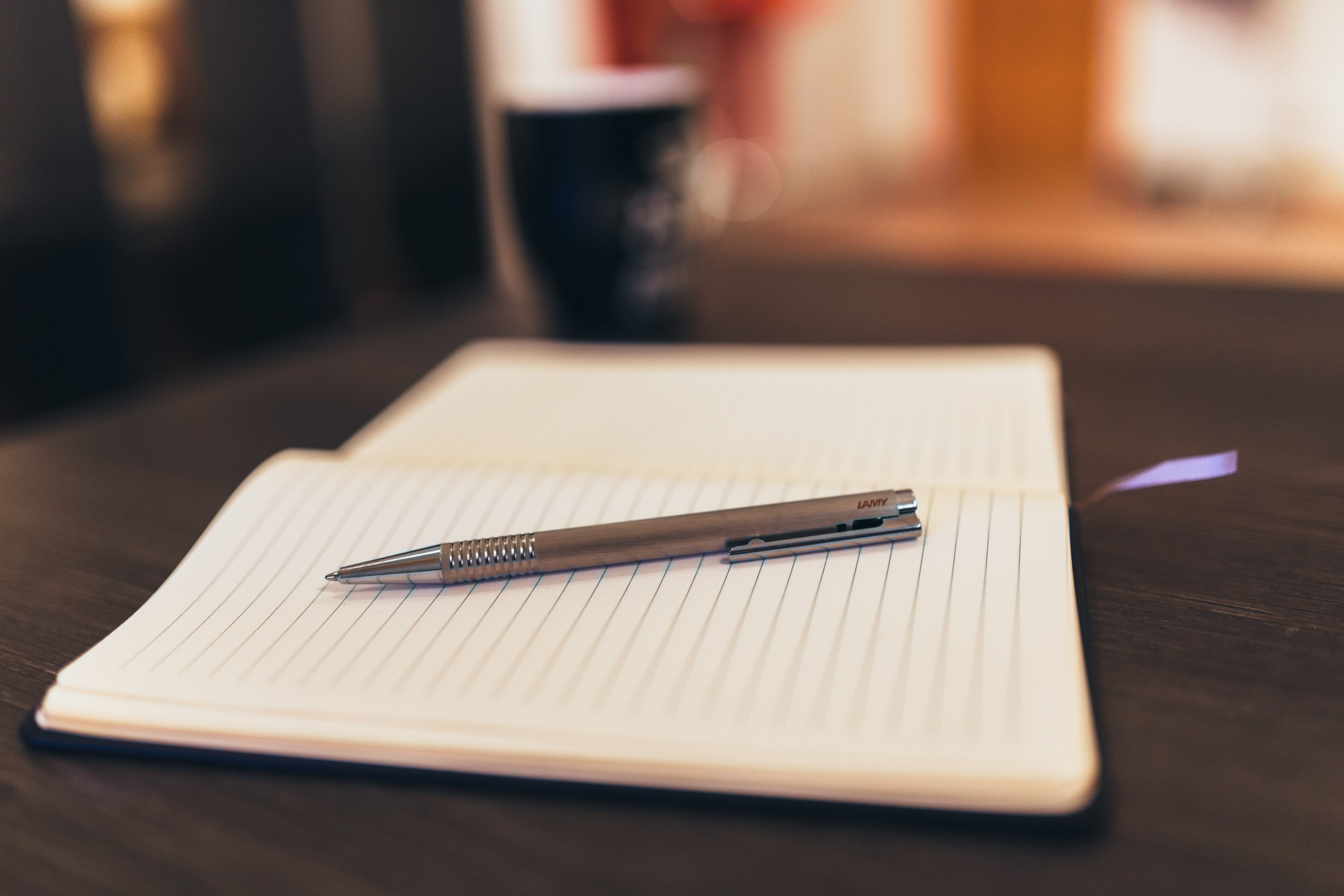 An open lined notebook with a pen resting on the page, a mug blurred in the background