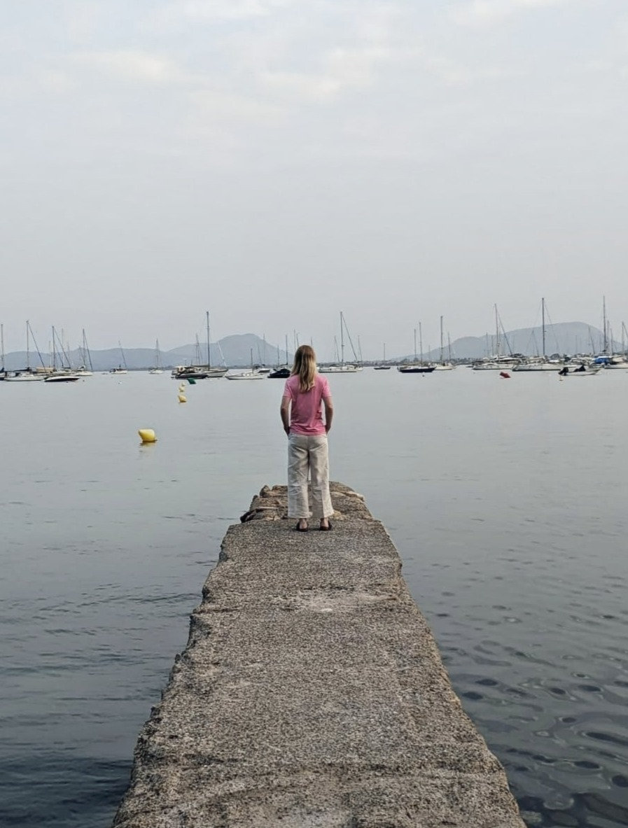 A woman standing at the end of a stone jetty, looking out across a harbour of moored boats