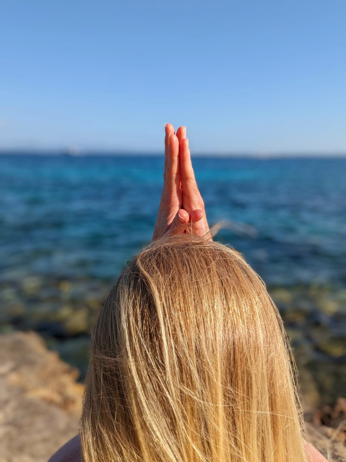 A woman seen from behind with her palms pressed together above her head, facing a bright blue sea