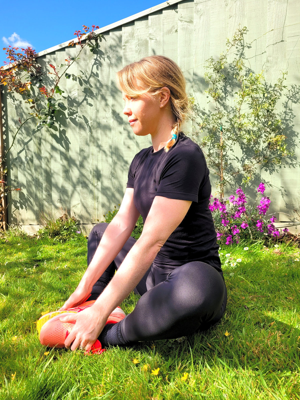 A woman in running kit sitting on the grass in a garden, stretching forward over one leg